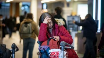 A frustrated passenger in a red parka rests their head in their hands and leans on a baggage cart at Vancouver International Airport.