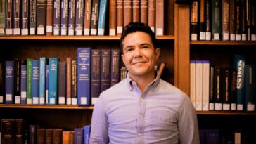 A smiling person stands in front of a bookcase.