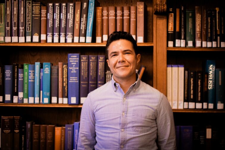 A smiling person stands in front of a bookcase.