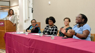 One woman at a podium and four women seated behind table in McBride Gathering Space